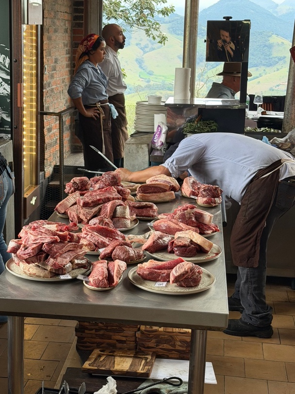 Bancadas com cortes de carne crua sendo preparadas para a desossa ao vivo ao lado do janelão com vista da serra