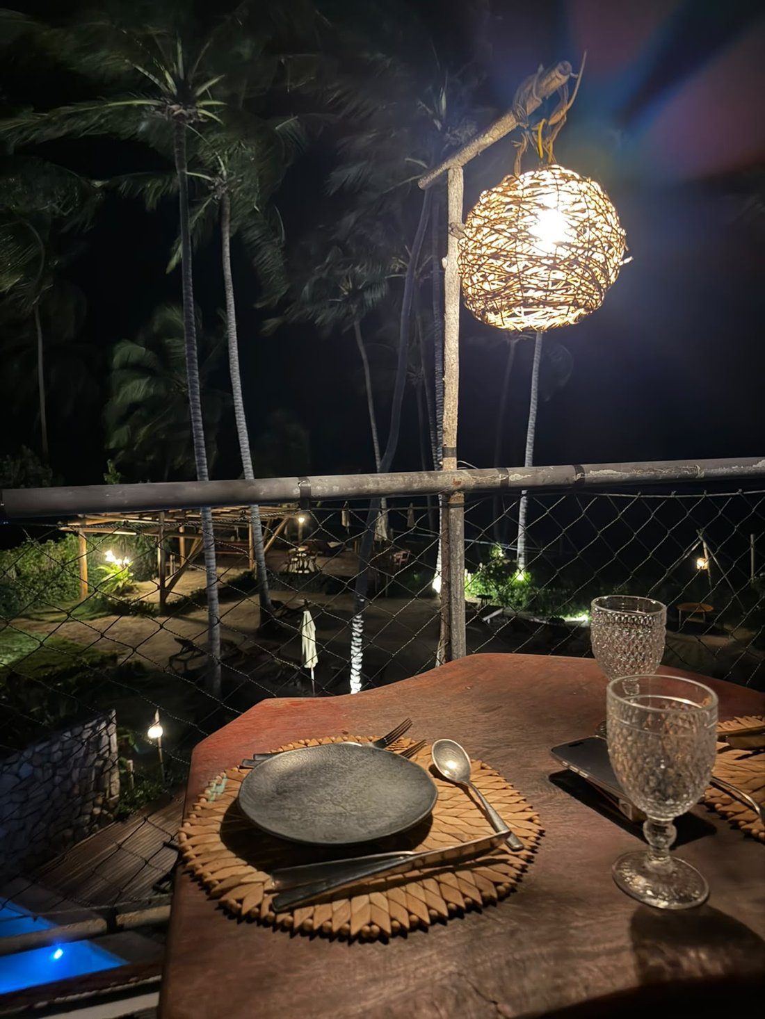 Jantar com vista à noite, sob luminária de fibra trançada, com as palmeiras iluminadas ao fundo